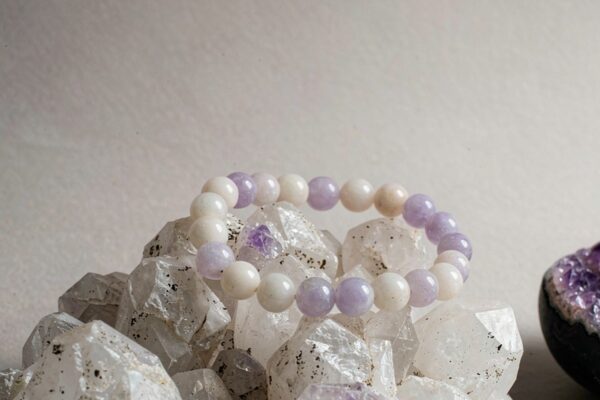 A lavender and white bead bracelet resting on a quartz crystal cluster beside a small amethyst geode
