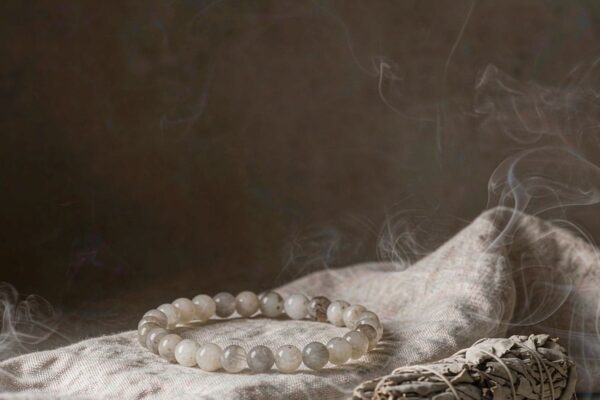 A crystal bead bracelet on linen surrounded by soft sage smoke with a dried sage bundle, illustrating how to cleanse a crystal bracelet with smoke