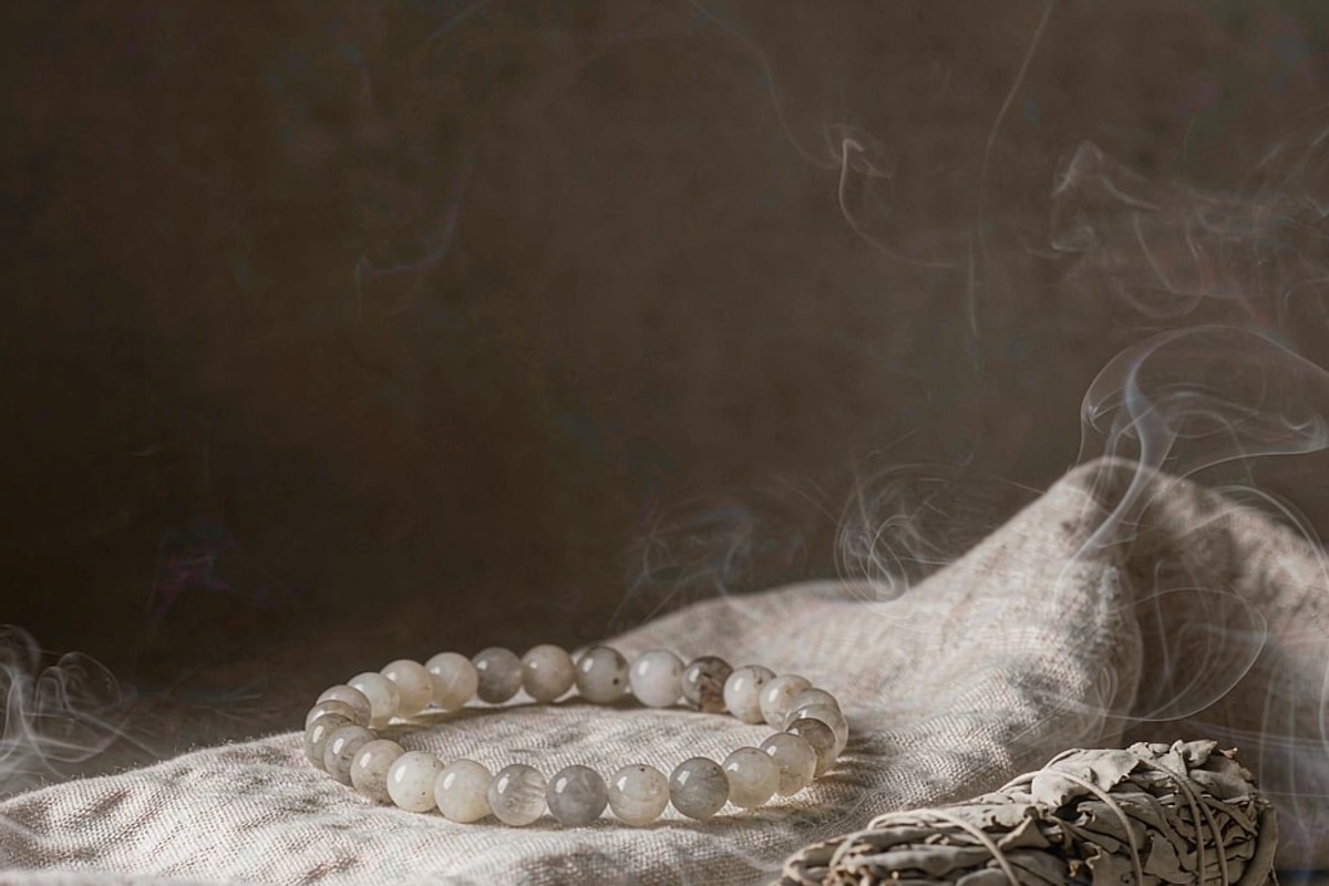 A crystal bead bracelet on linen surrounded by soft sage smoke with a dried sage bundle, illustrating how to cleanse a crystal bracelet with smoke