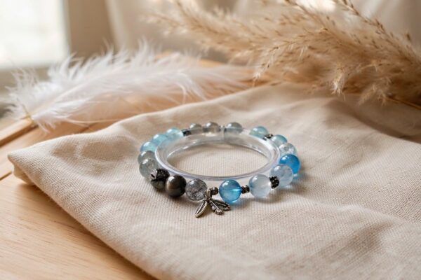 A grey rutilated quartz and icy blue bead bracelet with an antique silver leaf charm resting on ivory linen fabric on a light wooden table, with a white feather and pampas grass in the background