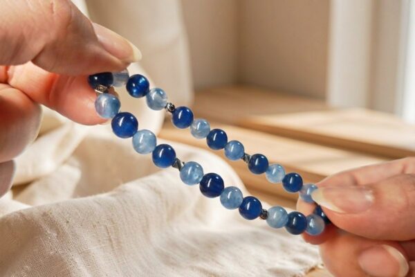 Hands holding a deep blue and light blue kyanite bead bracelet with silver spacers against a warm softly blurred interior with linen and wood