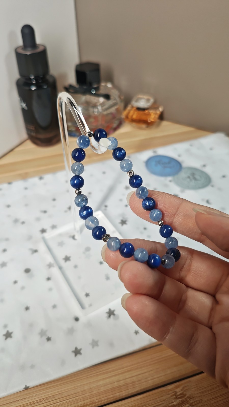Blue kyanite bracelet in deep blue and pale blue beads on a clear acrylic stand, held up beside blurred perfume bottles.