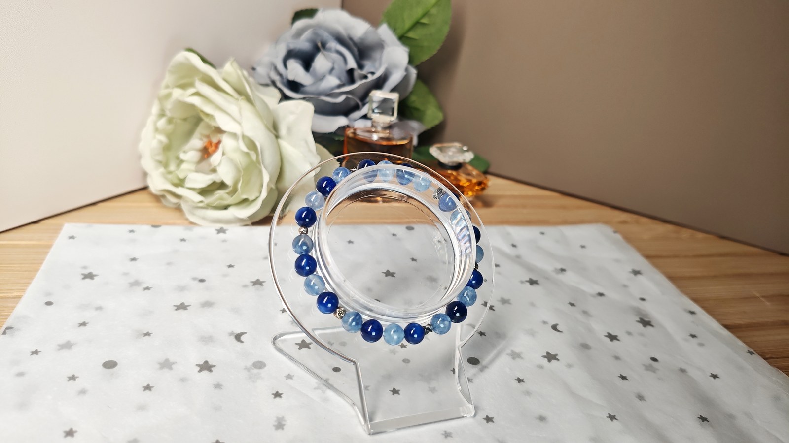 Two-tone blue kyanite bracelet resting on a round acrylic stand between cream and grey-blue roses.