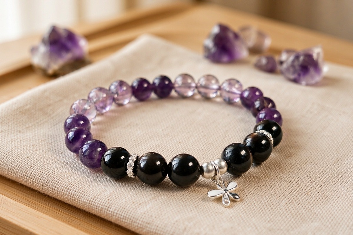 A purple fluorite and black obsidian bead bracelet with crystal spacers and an antique silver clover charm lying flat on ivory linen with raw amethyst crystals softly blurred in warm window light