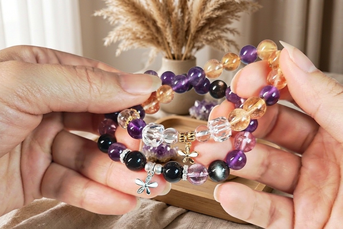 Two hands holding three stacked crystal bead bracelets of amethyst citrine and clear quartz with silver and gold flower charms against a softly blurred warm bohemian interior with pampas grass