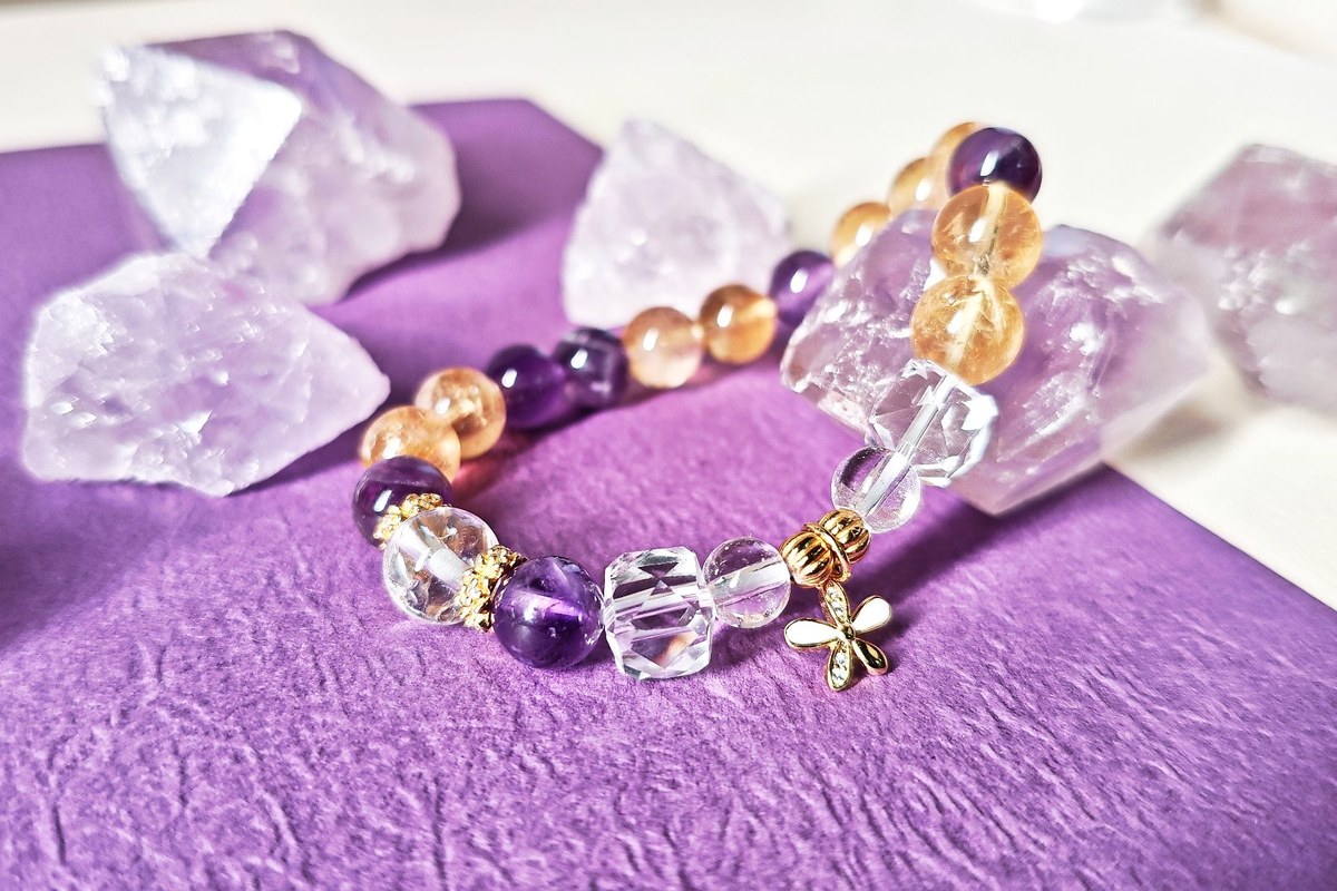 Amethyst, ice citrine, and clear quartz beads with a small gold flower charm, resting on purple fabric with clear quartz points.