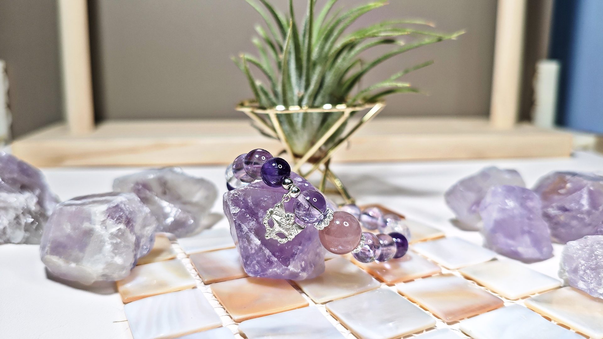 Amethyst crystal bracelet with a silver bead cap resting on a raw amethyst stone, beside an air plant on a tiled surface.