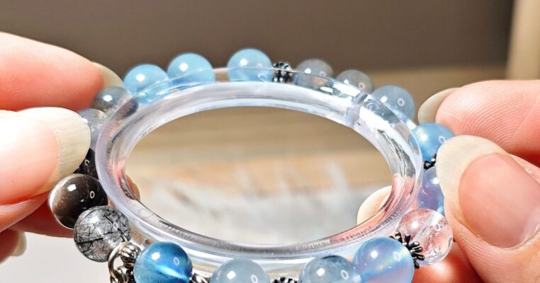Hands holding a handmade aquamarine bracelet with rutilated quartz beads and a silver feather charm