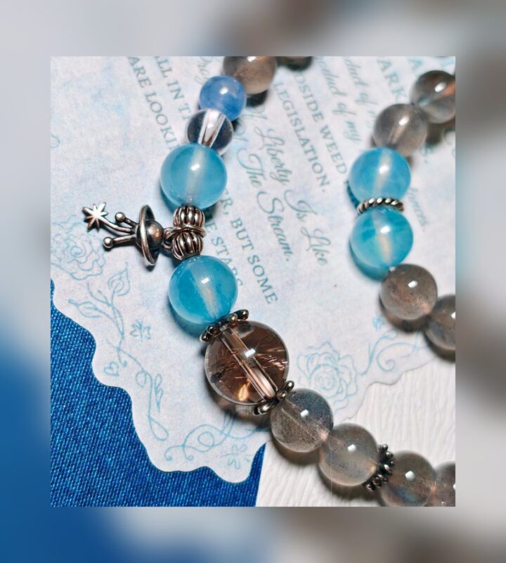 The Career Orbit crystal bracelet laid across a vintage printed page, showing blue aquamarine beads, grey moonstone and a silver planet charm.