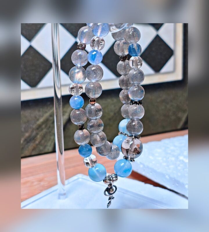 The Career Orbit crystal bracelet hanging on a silver display stand, showing its blue aquamarine, kyanite and black-gold quartz beads.