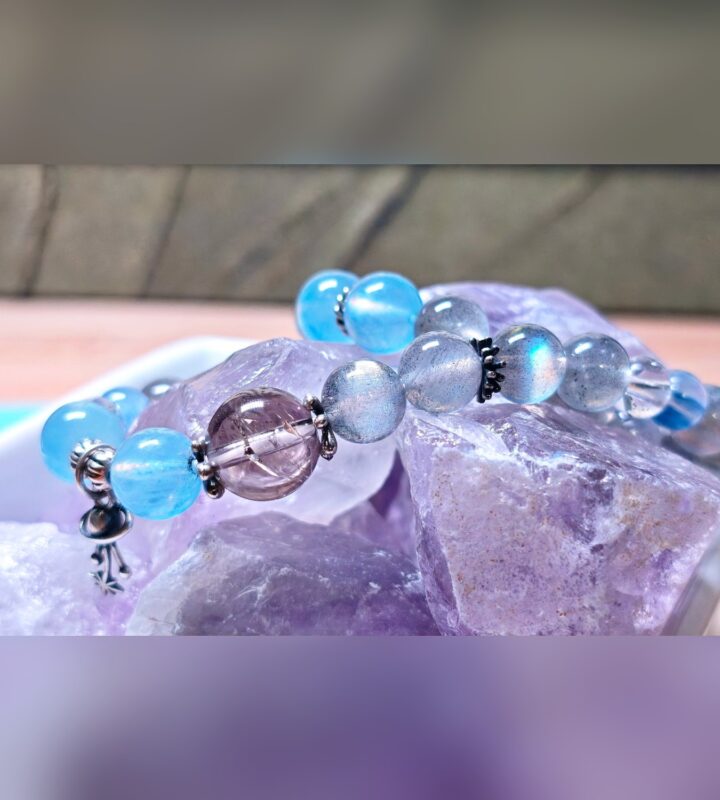 The Career Orbit crystal bracelet resting on a cluster of raw purple crystals, showing blue aquamarine and kyanite beads and a silver planet charm.