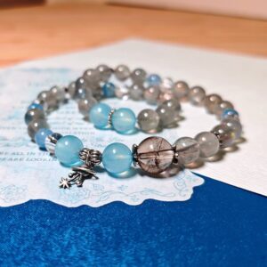 The Career Orbit crystal bracelet coiled on a printed page, with blue aquamarine and kyanite beads, dark black-gold quartz beads, and a small silver planet charm.