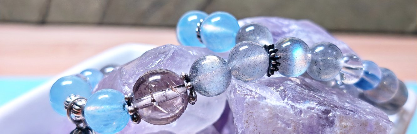 Close-up of a crystal bracelet for women in aquamarine, moonstone and smoky quartz resting on raw amethyst stone