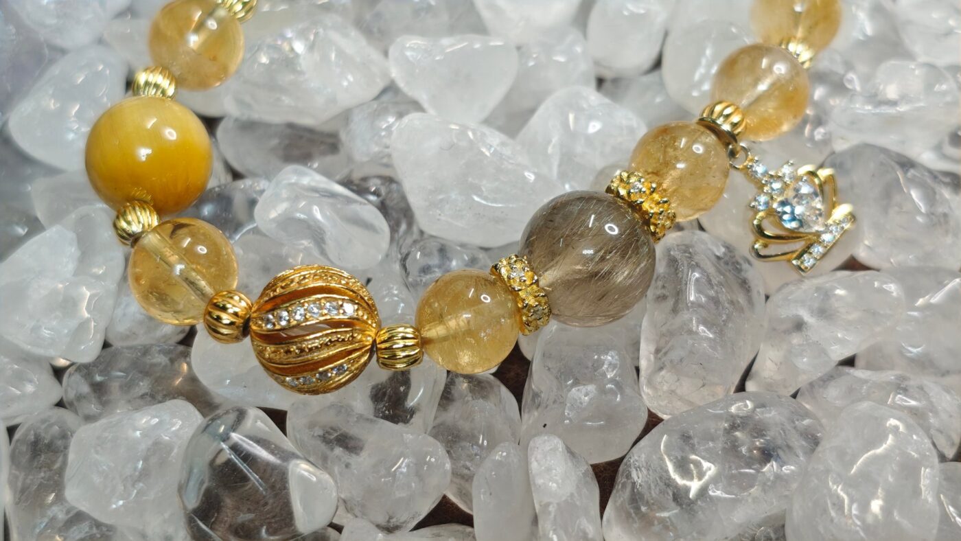 Close-up of the golden rutilated quartz, ice citrine and golden tiger's eye beads with gold-tone spacers and a gold crown charm, resting on clear quartz crystals.