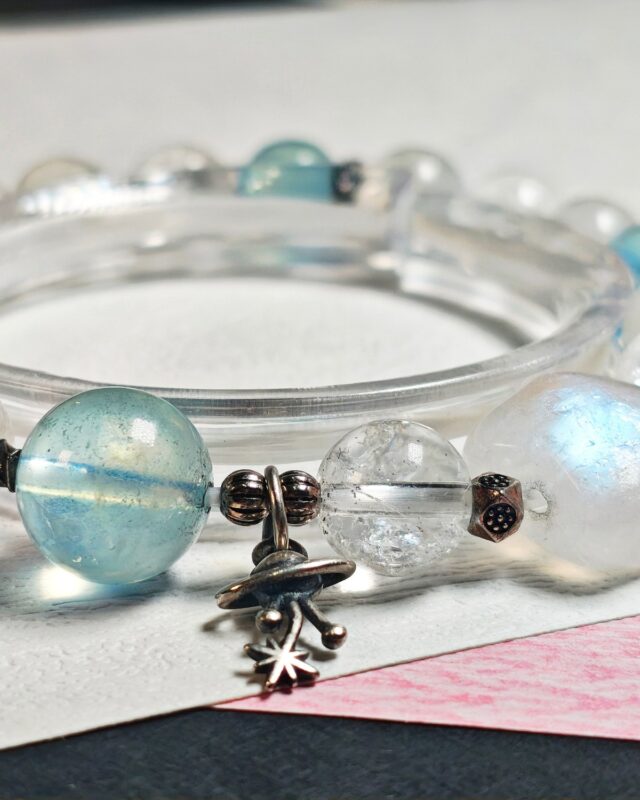 Close view of the s925 sterling silver planet charm beside an aquamarine bead, a clear quartz bead and a moonstone, with spacer beads
