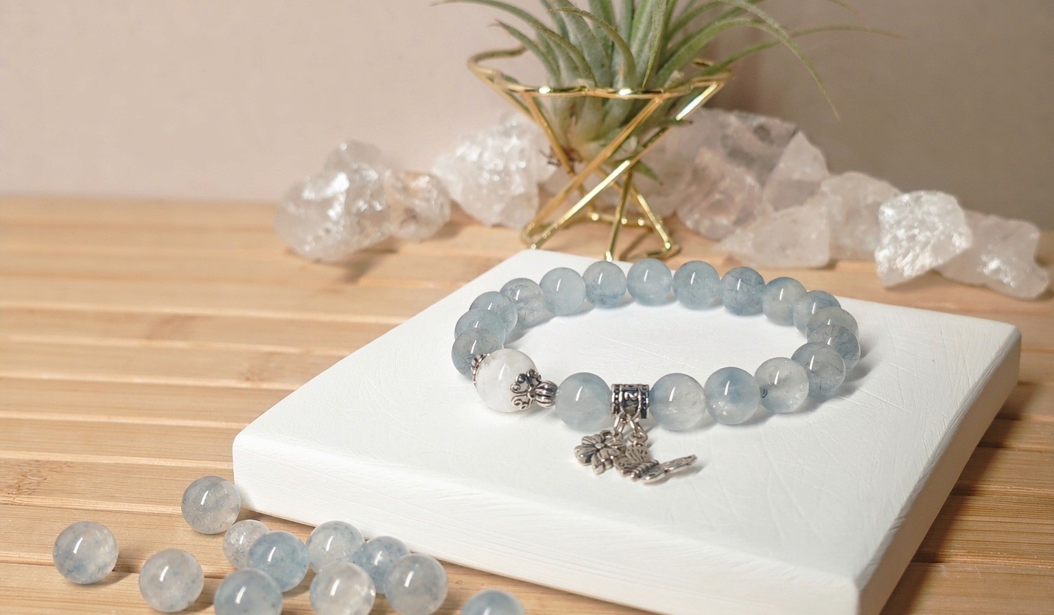 Phantom quartz bracelet with pale blue rutilated beads and one white phantom bead on a white display block, crystal points behind