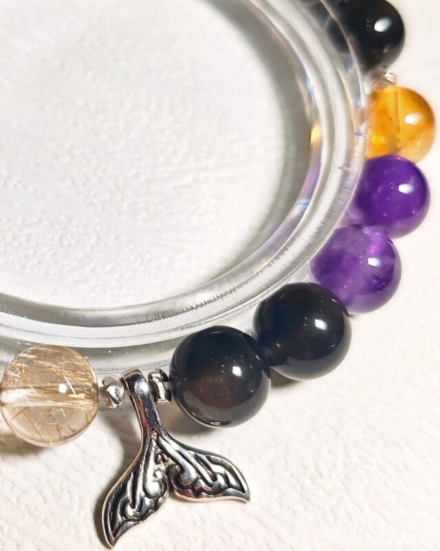 Close view of the Rising Current crystal bracelet on a clear display stand, with its amethyst, Ice Citrine and golden rutilated quartz beads and mermaid tail charm in sharp detail.