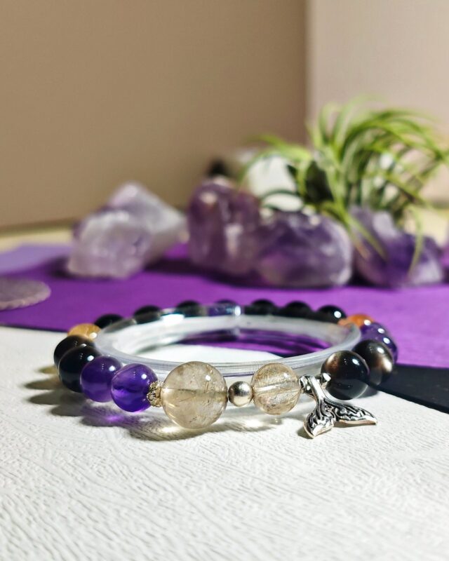 Rising Current crystal bracelet on a clear display stand over a purple cloth, with an amethyst cluster and a green plant behind it.