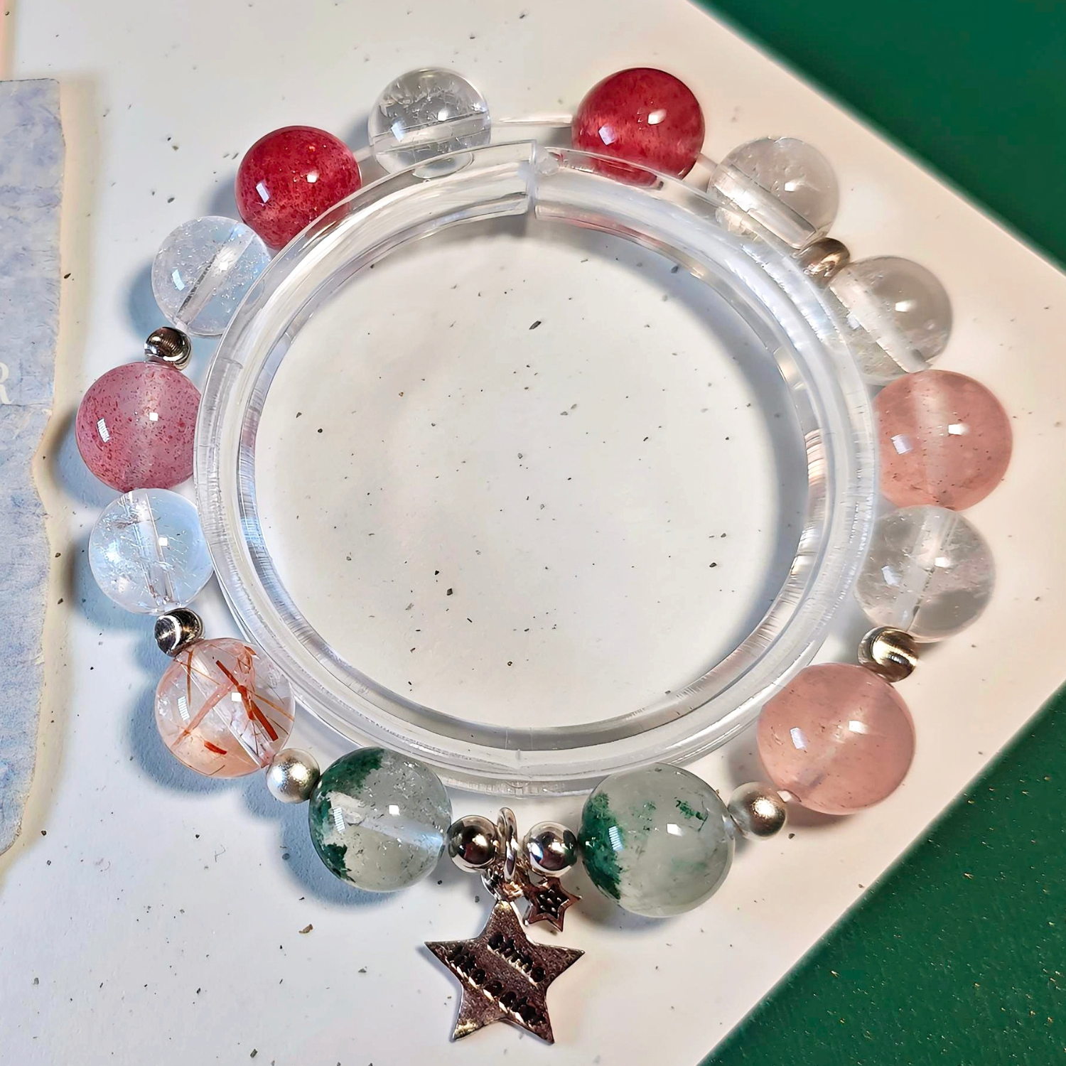 Black gold super seven bracelet on a clear display stand, with green phantom quartz, ice strawberry quartz and clear quartz beads.