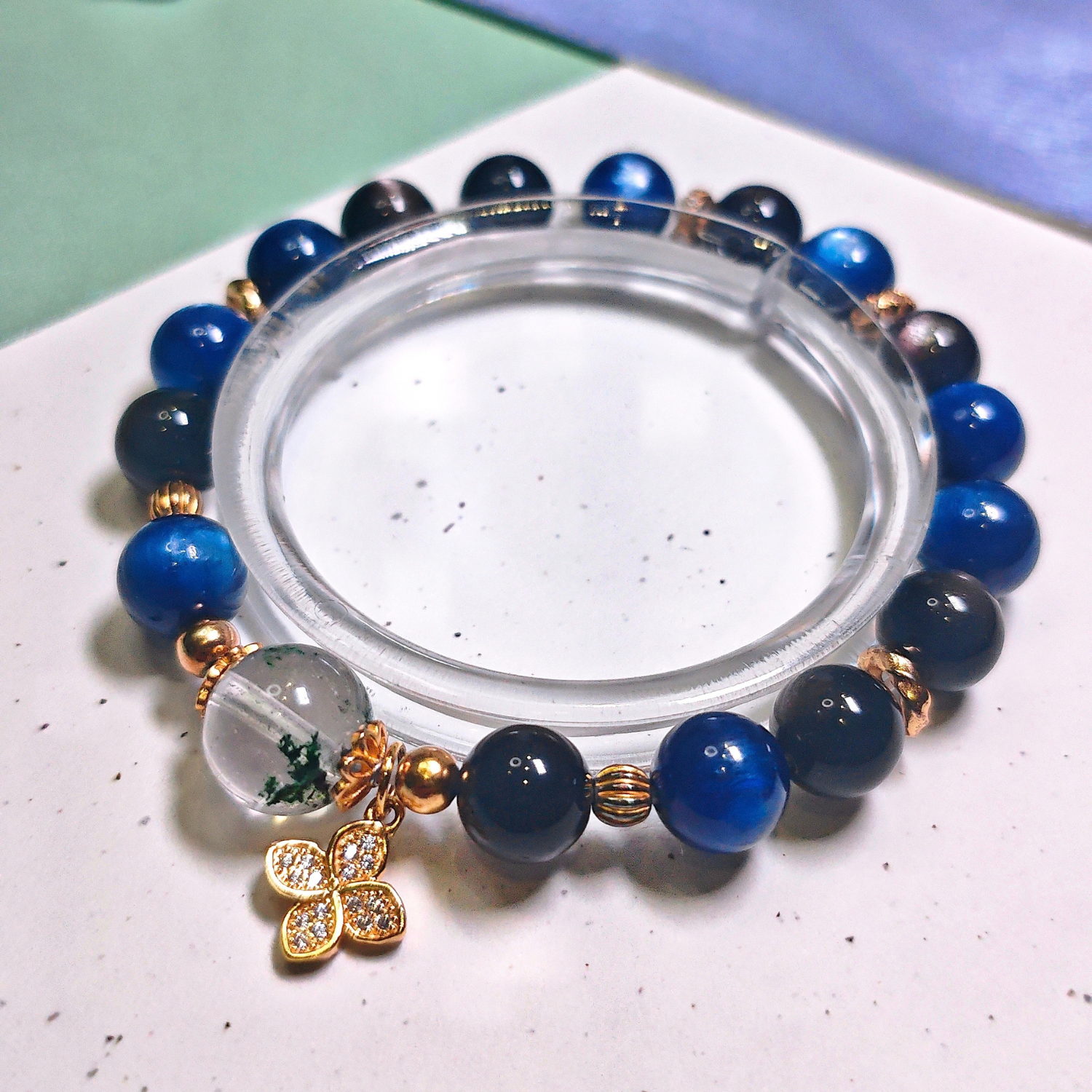 Green phantom quartz bracelet on a clear display stand, with mossy green phantom quartz, deep blue kyanite and silver obsidian beads.
