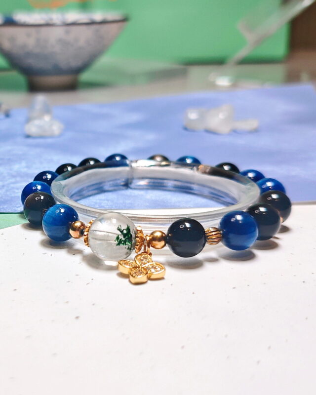 Steady Presence crystal bracelet on a clear display stand over a pale cloth, with soft florals and a ceramic bowl behind it.