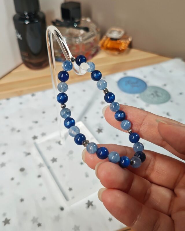 Still blue crystal bracelet on a clear stand beside perfume bottles, deep blue and pale blue beads alternating