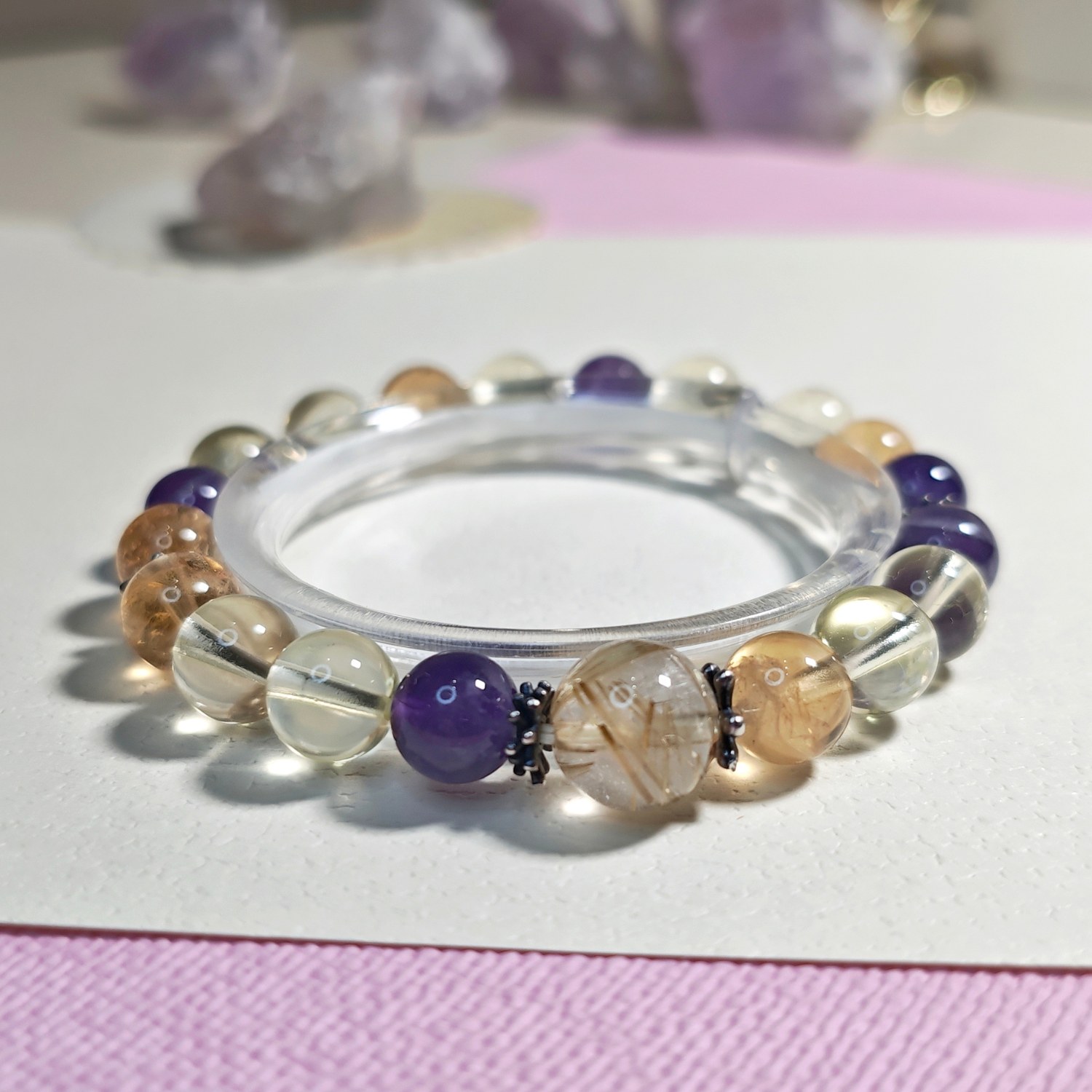 Golden rutilated quartz amethyst bracelet standing on a pink surface with an amethyst cluster behind
